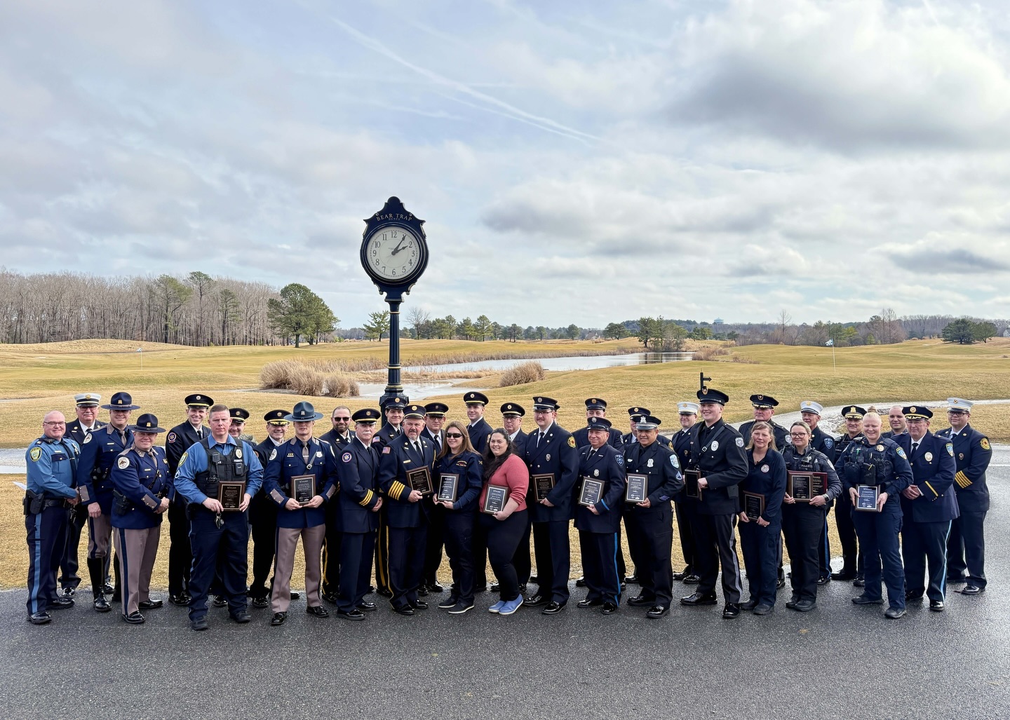 A large group of uniformed police officers and emergency personnel stand outdoors on a paved path at the 2026 Joshua M. Freeman First Responder Awards at Bear Trap Dunes. Many of them are holding award plaques. They are posed in a line beneath a tall decorative street clock that reads “Bear Trap.” Behind them are rolling grassy fairways, a small pond, scattered trees, and a cloudy sky.